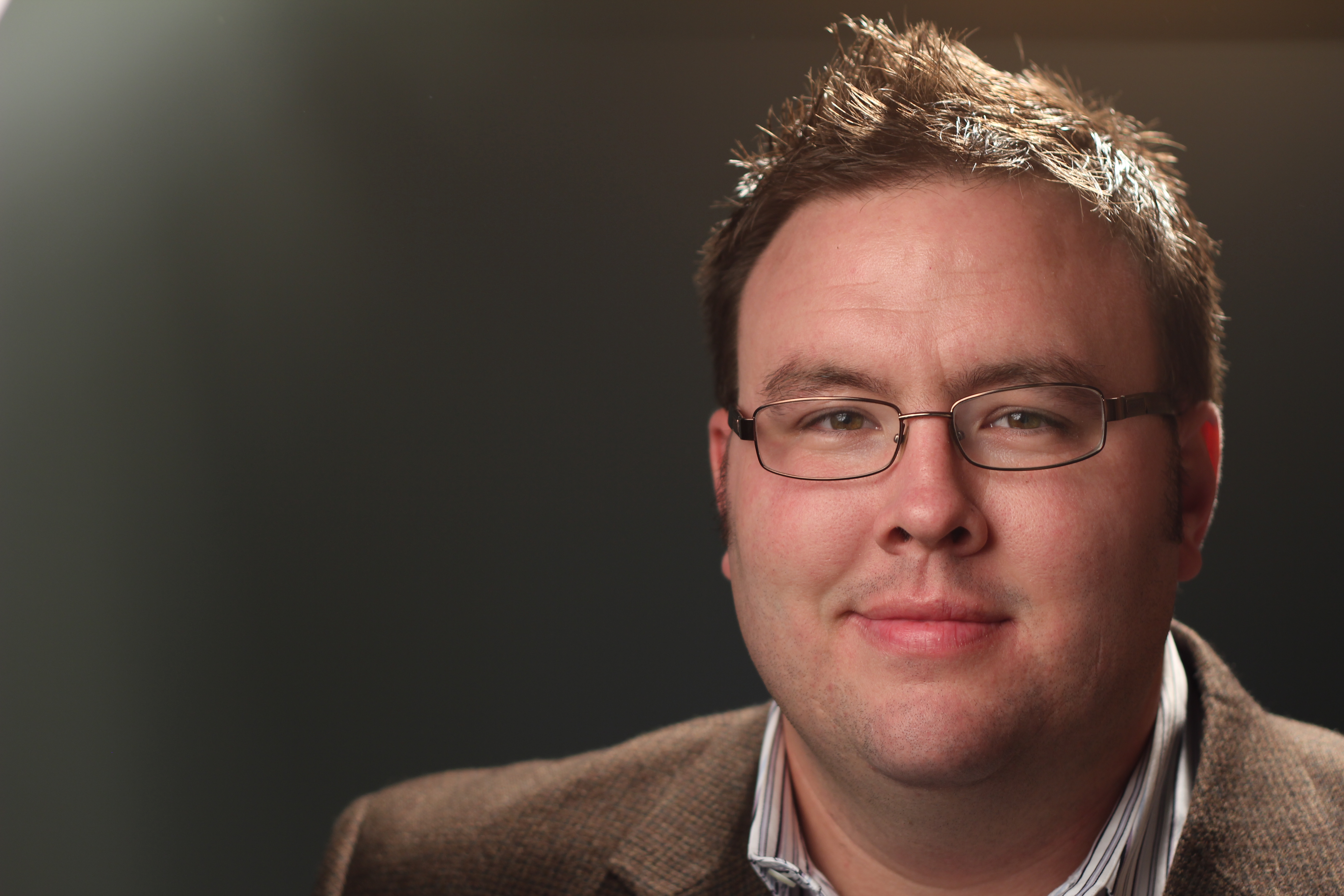 Portrait of a man wearing glasses and a brown jacket against a dark background.