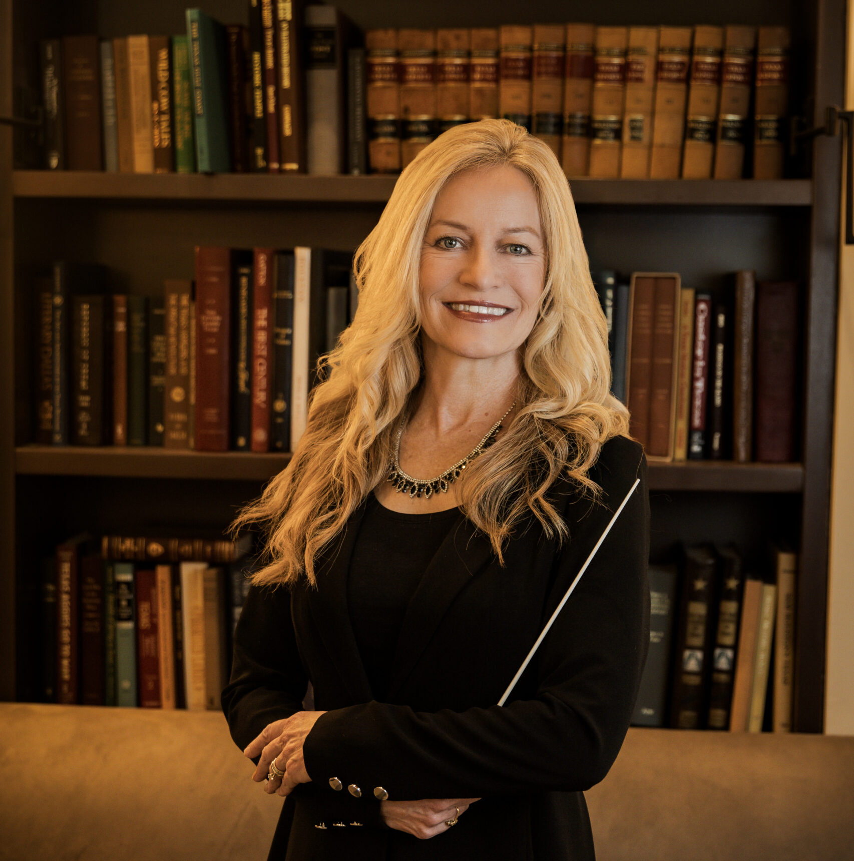 Smiling woman with long blonde hair holding a conductor's baton in a library.