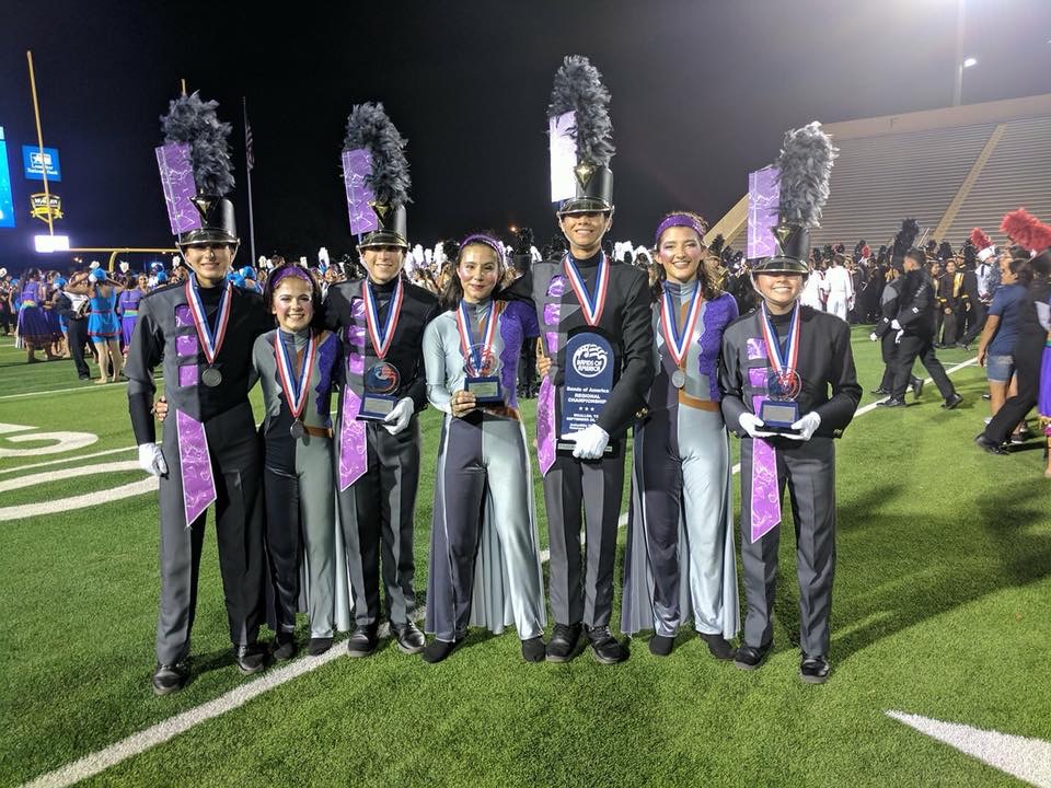Marching band members in purple uniforms posing with trophies on a field at night.