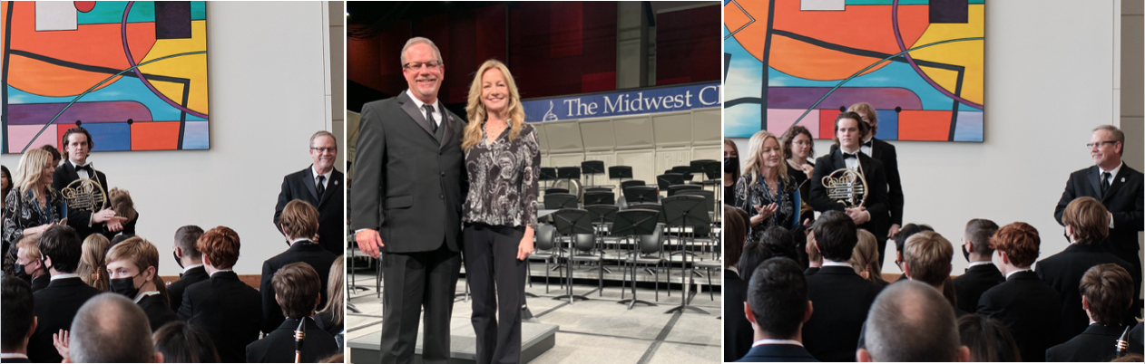 Two individuals stand smiling on a stage with chairs behind them.